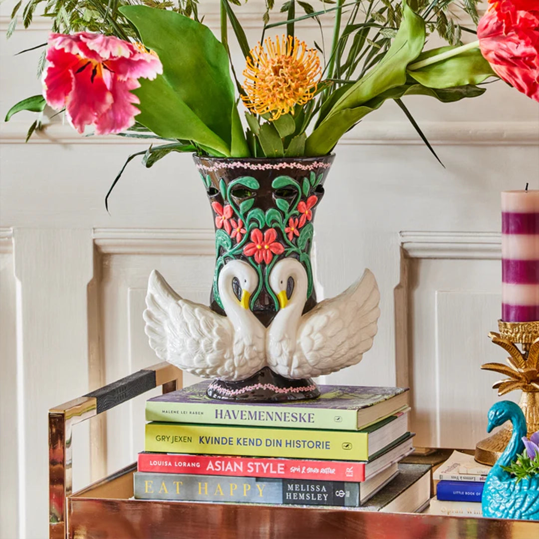Vase en céramique noir avec des cygnes blancs, posé sur une pile de livres, décoré avec des fleurs exotiques.