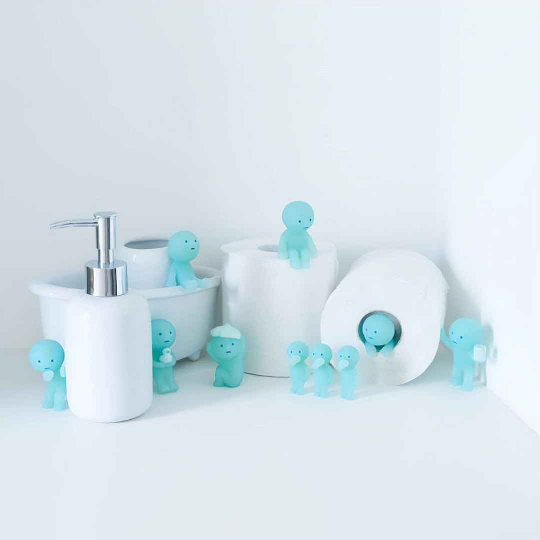Figurines Smiski de la Série Toilettes, disposées autour d'articles de toilette tels que des rouleaux de papier hygiénique et un distributeur de savon, brillent dans l'obscurité.